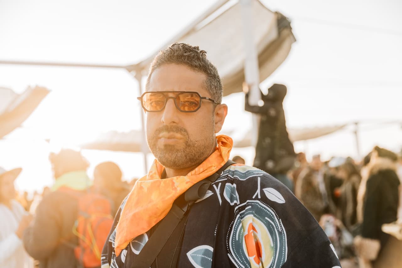 A stylish man with a beard and orange-tinted sunglasses looks directly at the camera, wearing a patterned jacket and an orange bandana in a