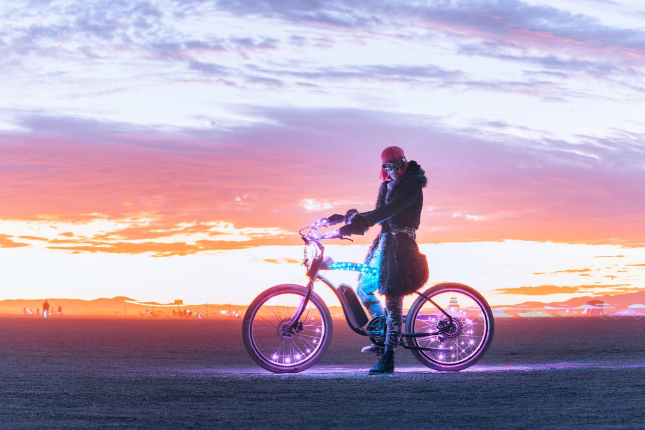 A person on a glowing electric bicycle stands silhouetted against a vibrant orange and purple sunset sky in an open, flat landscape.