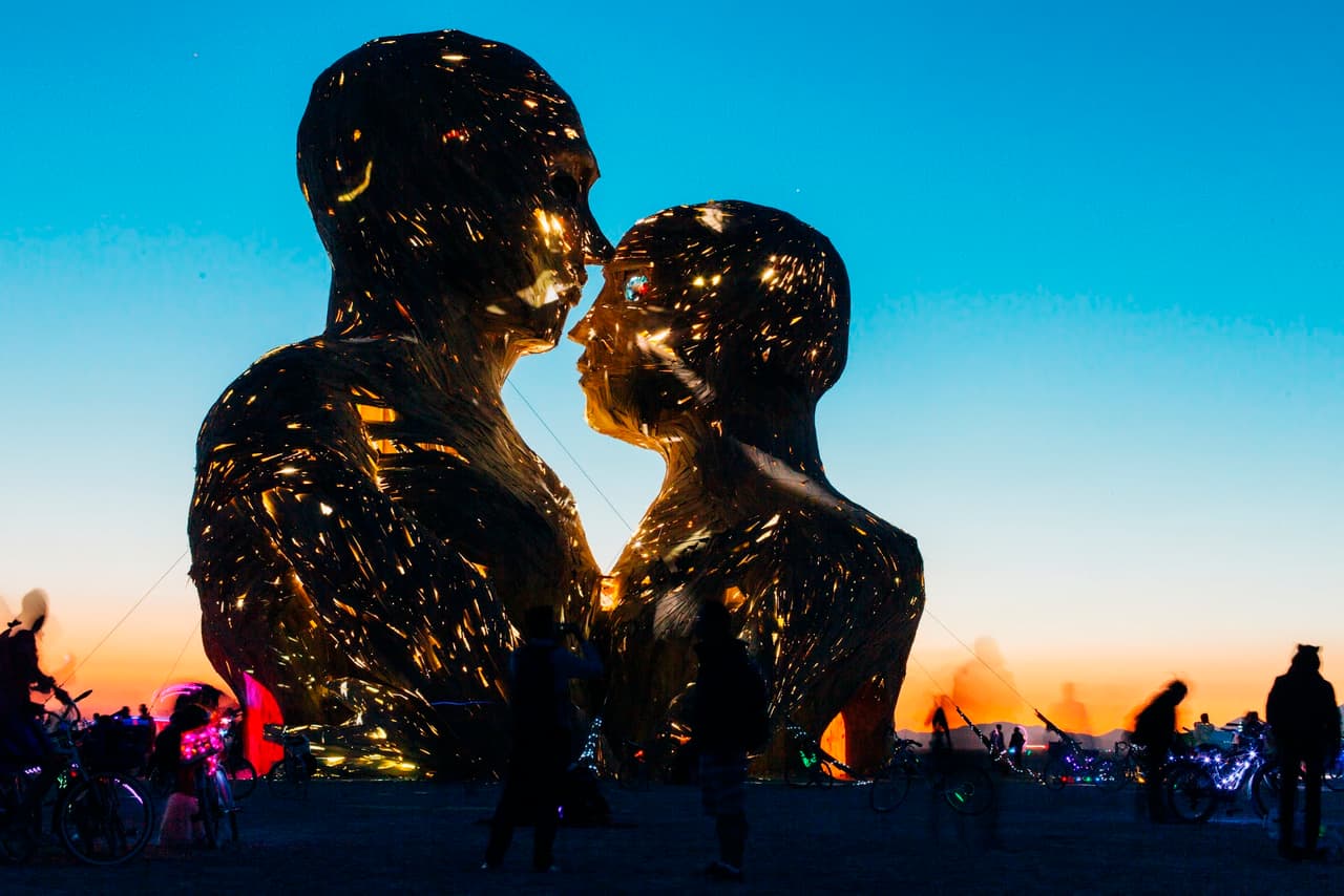 Two colossal golden human figures, illuminated from within, face each other under a vibrant sunset sky, surrounded by silhouetted onlookers.