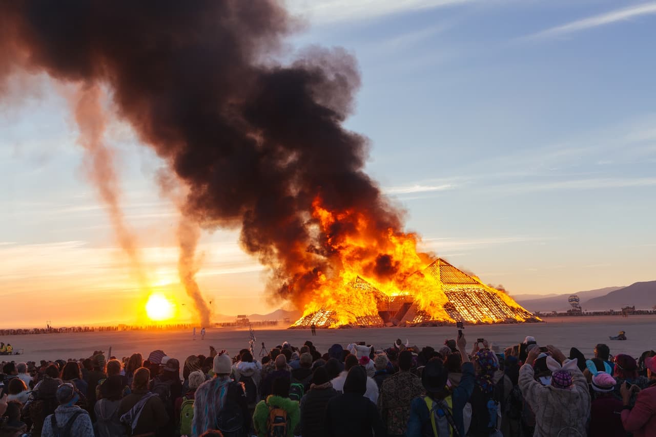 A massive wooden pyramid-like structure burns fiercely against a sunset sky, watched by a large crowd in a desert landscape.