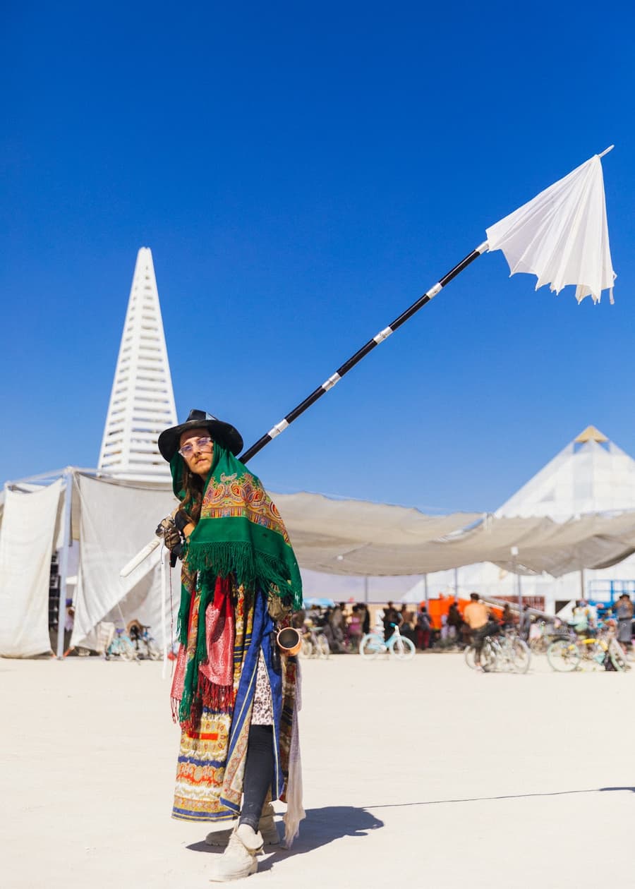 A person in a colorful, layered outfit and hat carries a white flag on a pole across a desert landscape under a clear blue sky.