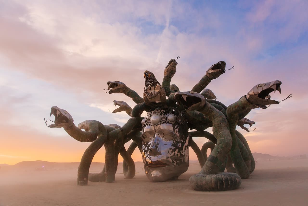 A large, metallic sculpture resembling a Medusa-like head with numerous snake bodies for hair stands prominently in a vast desert landscape