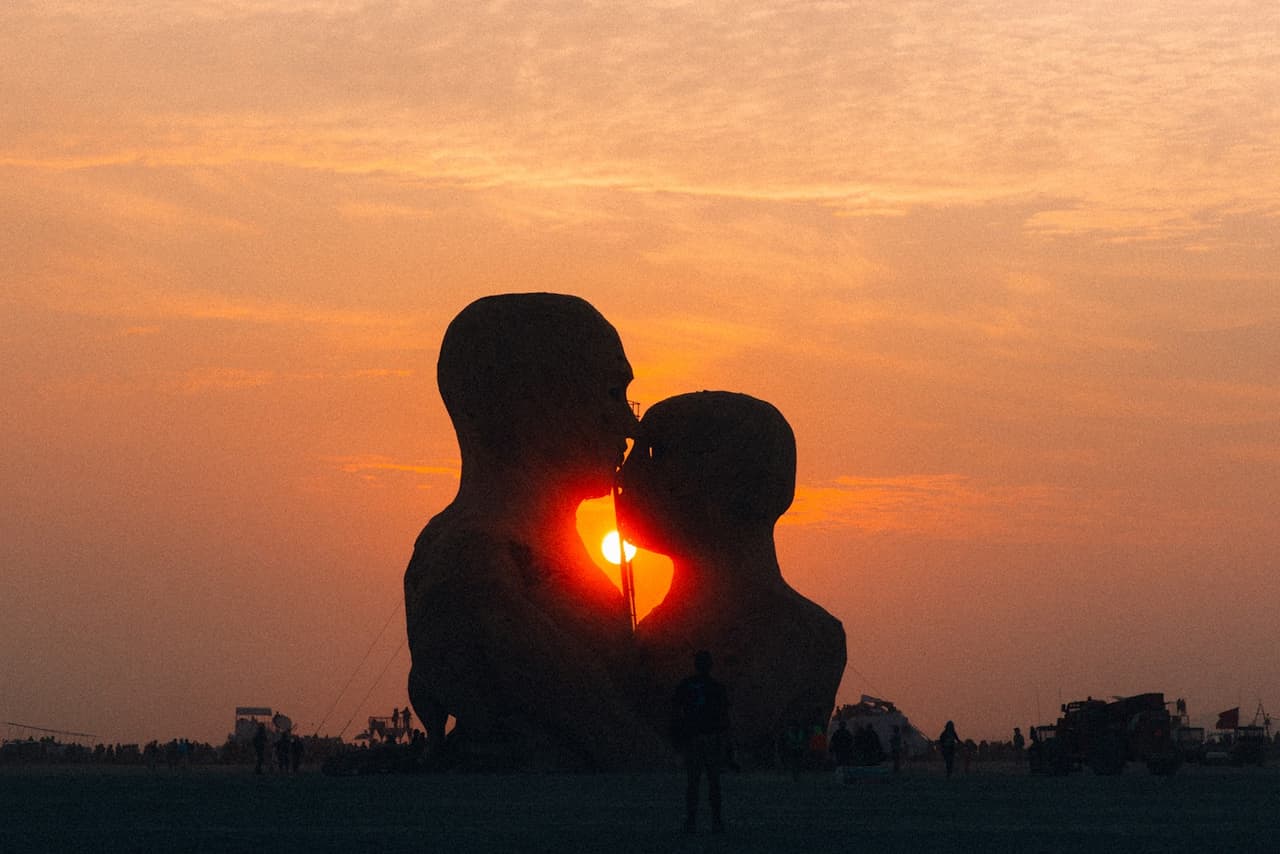 Two massive human-like sculptures are silhouetted against a vibrant orange sunset, appearing to share a tender moment.