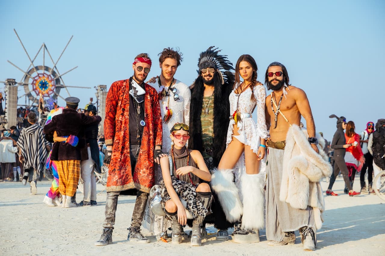 A diverse group of seven people in elaborate, bohemian-inspired festival attire poses confidently in a sunny desert landscape.