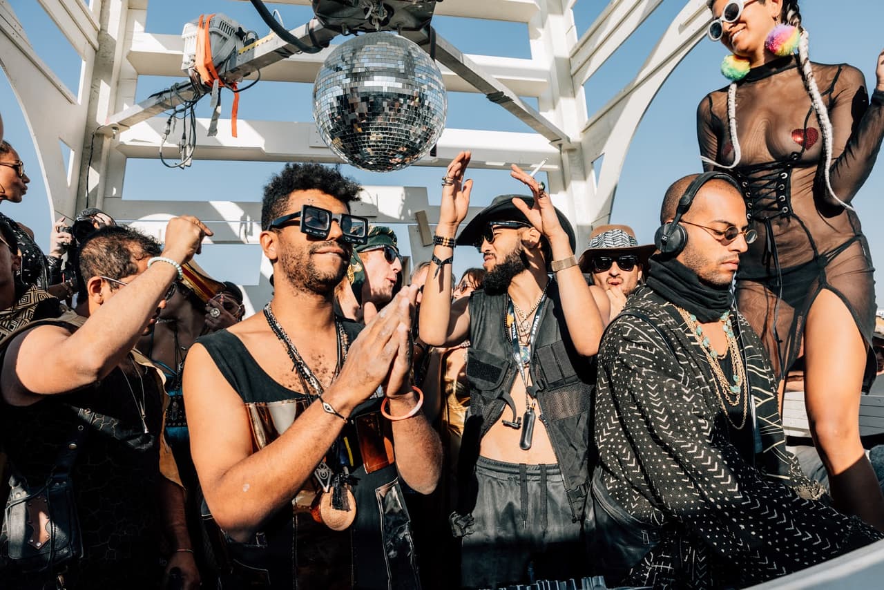 A diverse group of people in elaborate outfits dance and celebrate under a bright sky with a disco ball at an energetic outdoor event.
