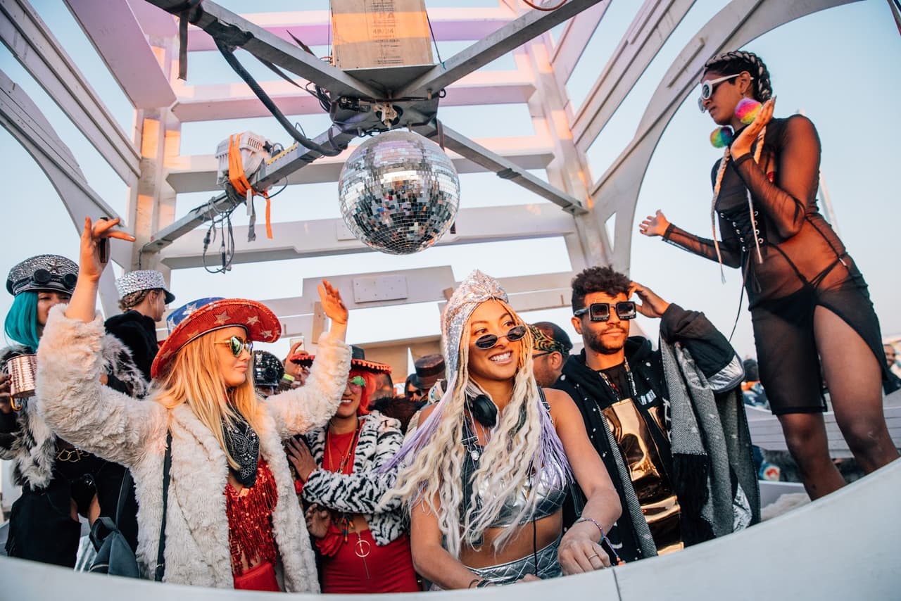A vibrant outdoor festival scene shows a smiling DJ surrounded by energetic partygoers under a large disco ball.
