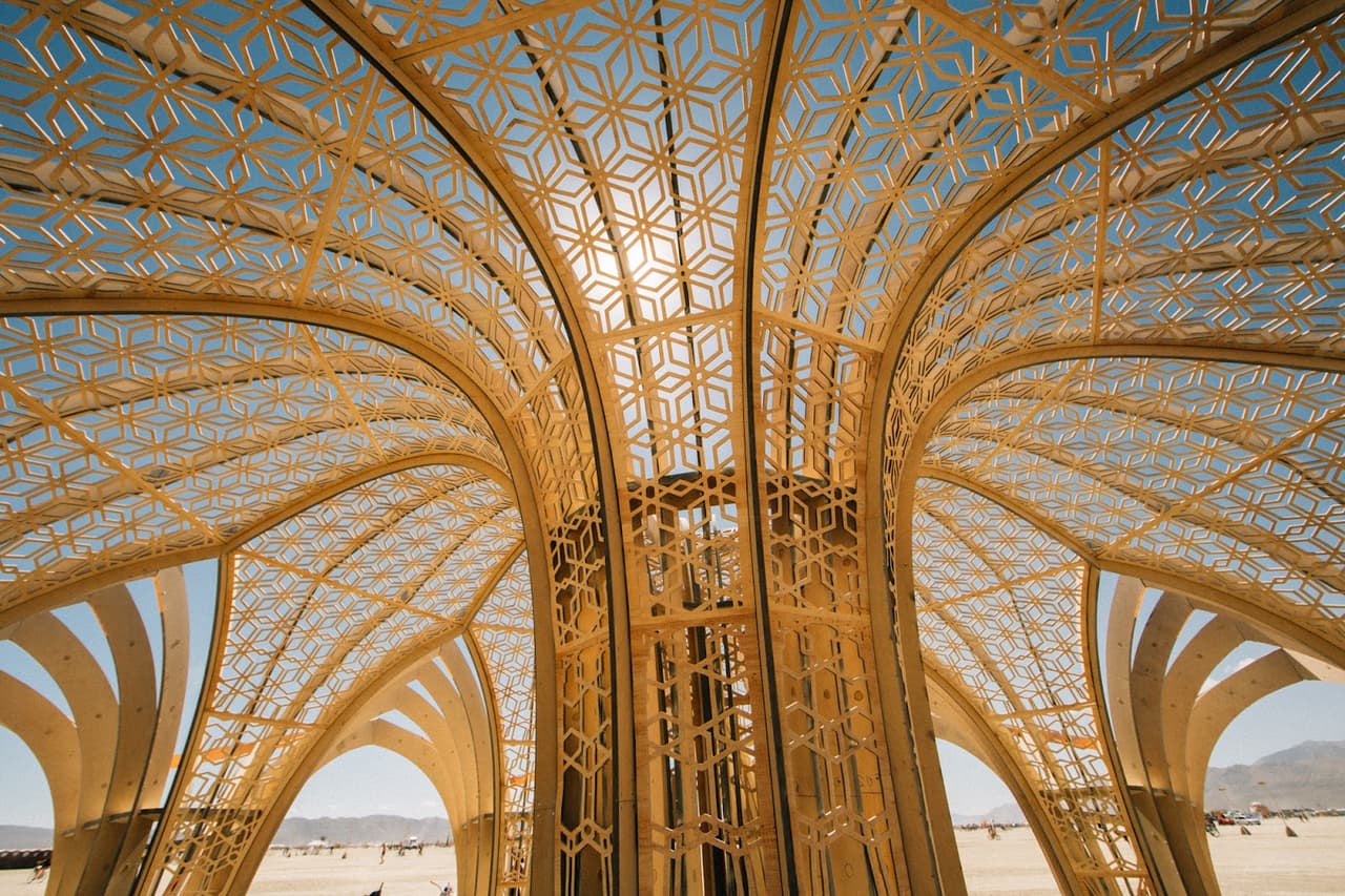 A magnificent golden structure with intricate geometric patterns arches towards a bright blue sky, with the sun peeking through its lattice.