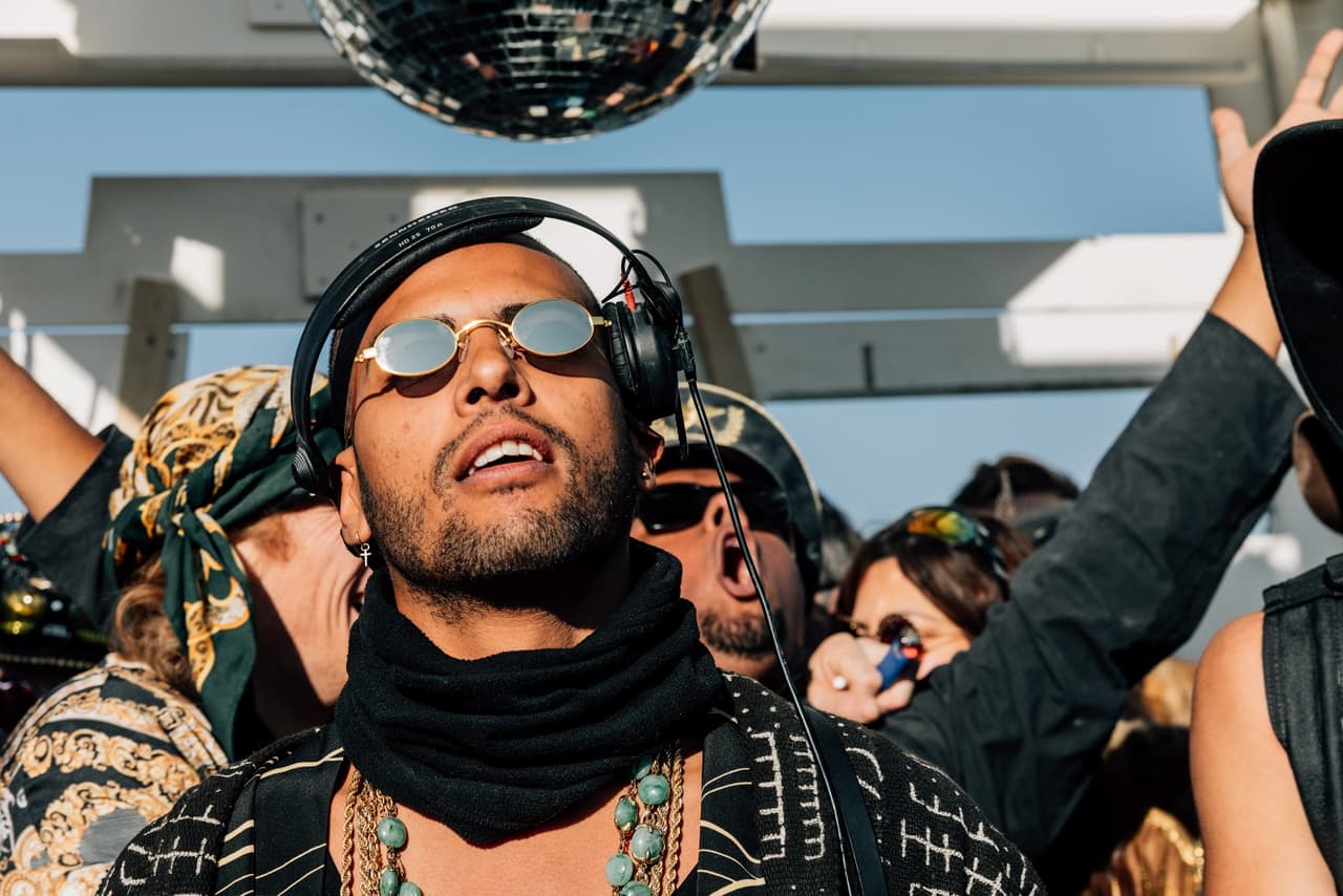 A man with sunglasses and headphones looks up intently at a disco ball, surrounded by a lively crowd under a bright sky.