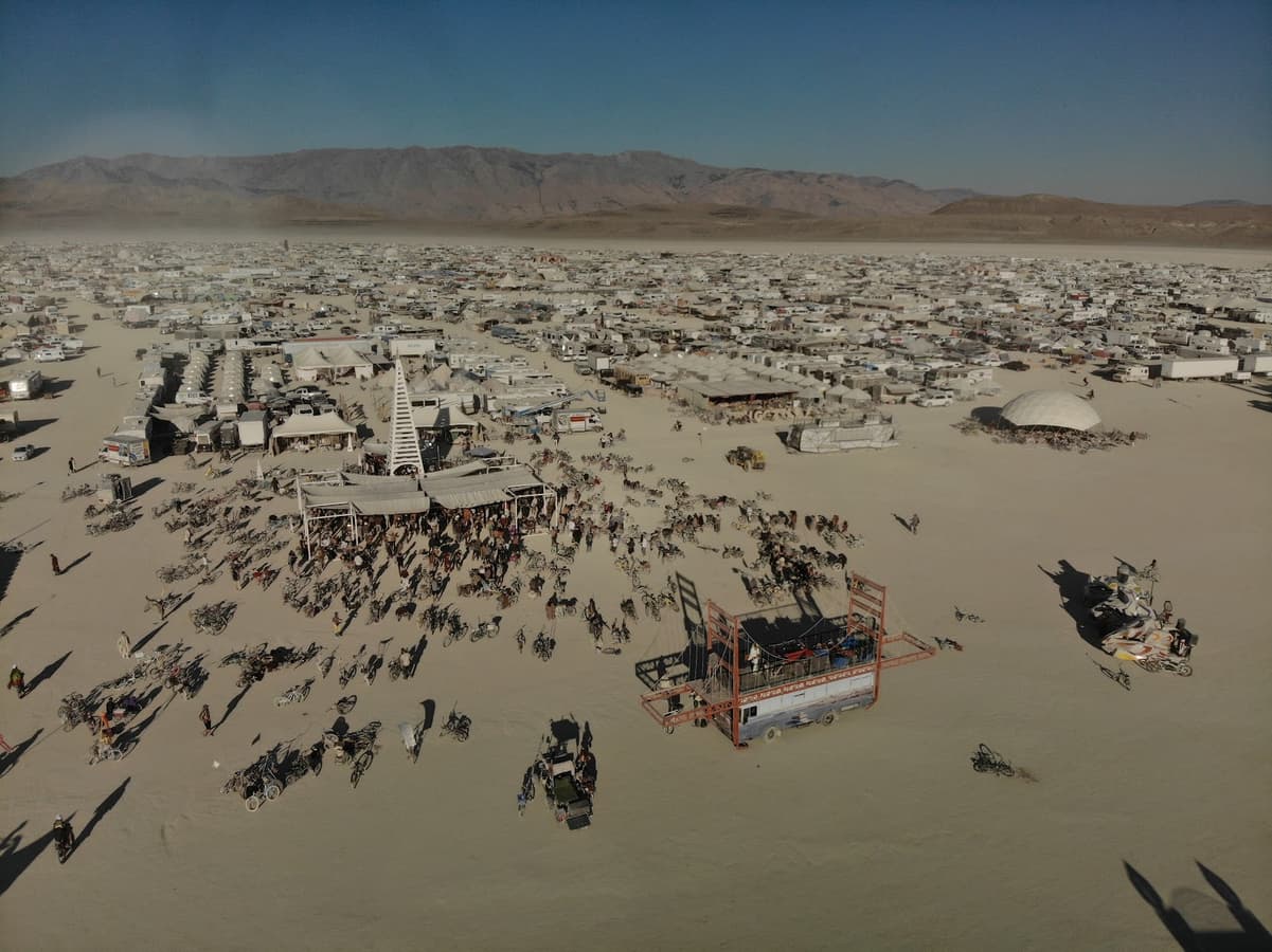 An expansive aerial view captures a temporary desert city bustling with people and unique structures under a clear blue sky, with mountains