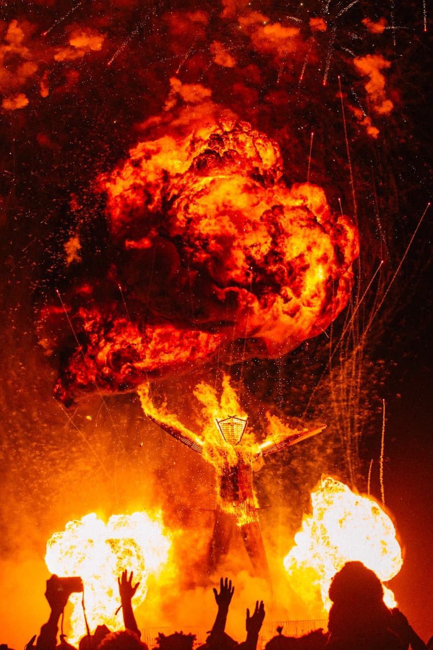 A performer in a golden costume stands amidst a massive fiery explosion, holding a giant fireball above their head while smaller flames erup