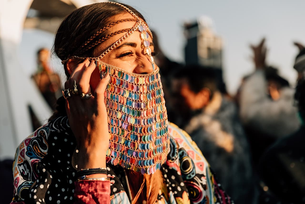 A woman with a vibrant, multi-colored beaded face veil and intricate head jewelry smiles, bathed in golden hour sunlight.