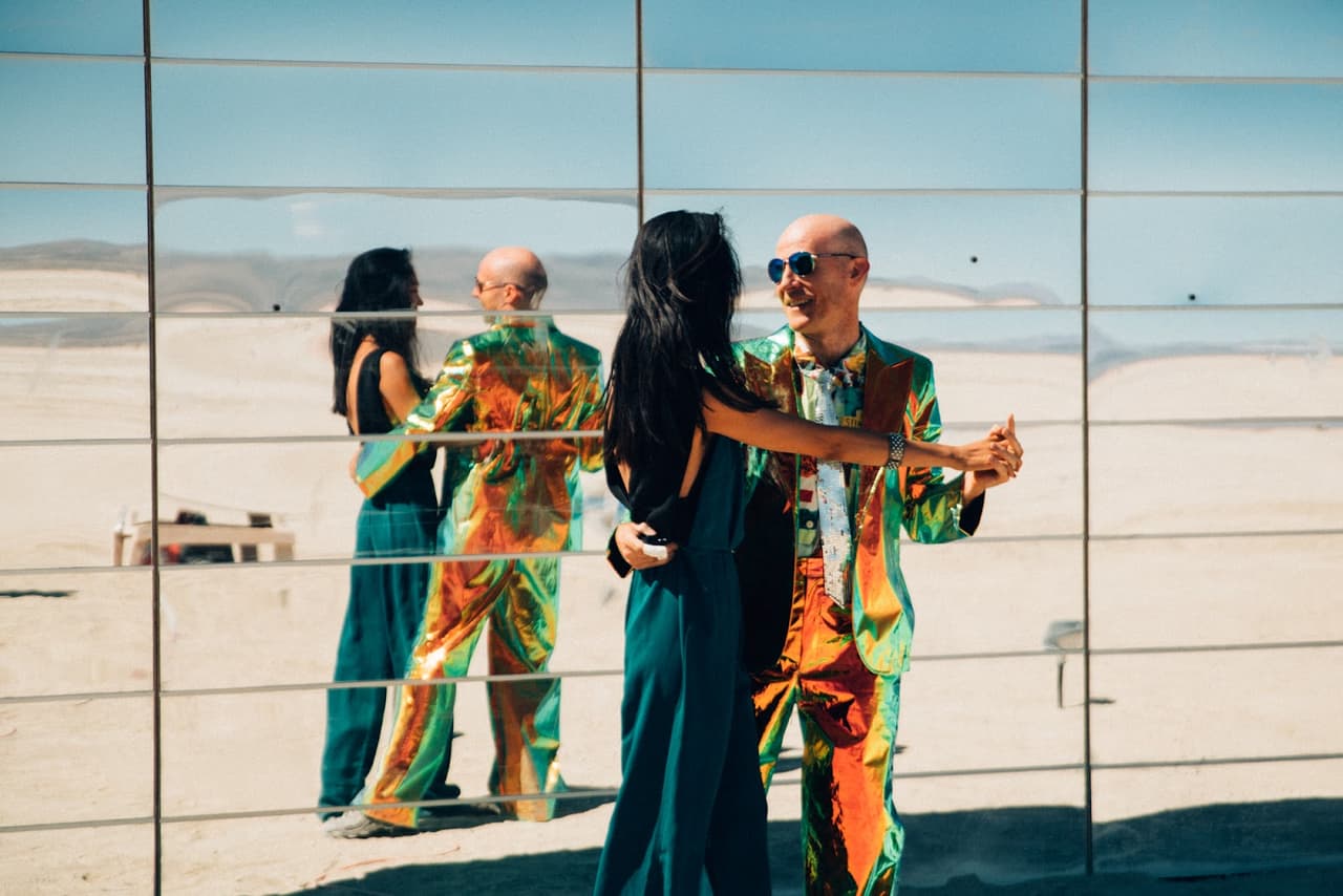 A couple in vibrant, reflective outfits dances in front of a large mirror wall art installation in a vast desert landscape under a clear blu