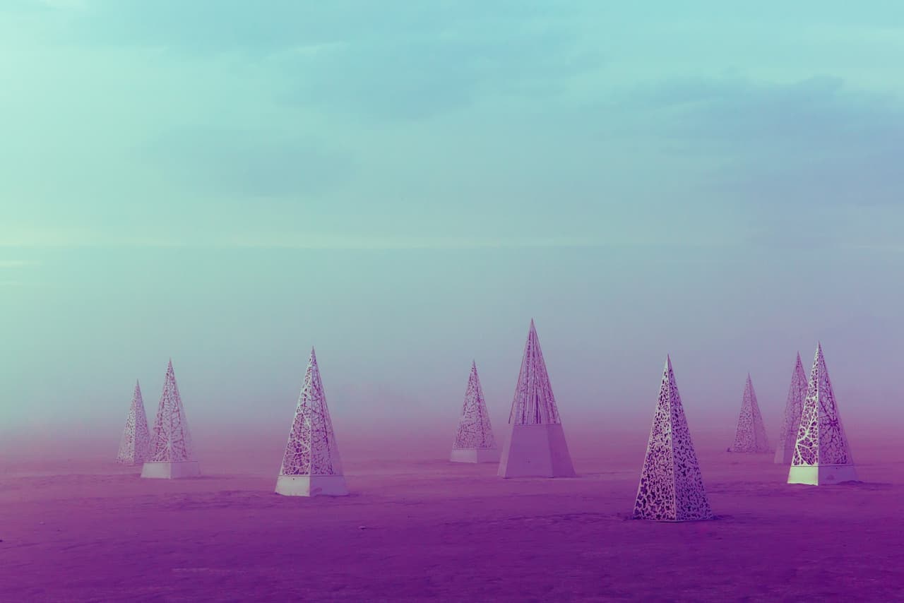 Numerous intricately patterned conical structures emerge from a vibrant magenta landscape under a turquoise sky, shrouded in a mysterious fo